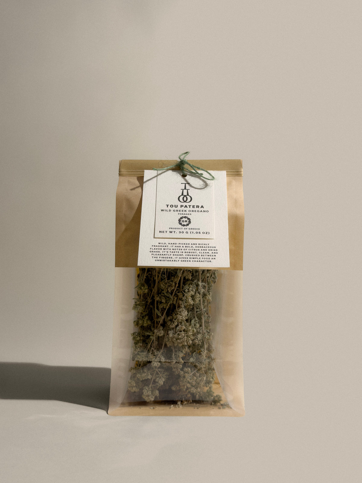 A clear plastic pouch of dried tea leaves with a kraft paper top band and a white paper label, tied with a thin string, set against a plain light gray background.