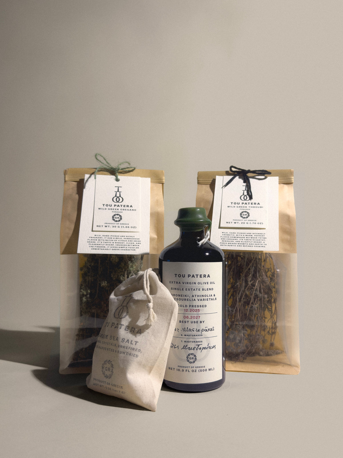 A set of tea products from Tou Patera  arranged on a neutral studio background. The set includes two clear bags of loose leaf tea with kraft paper sleeves and white labels, a small white drawstring pouch of sea salt, and a dark glass bottle of herbal tea with a green cap.