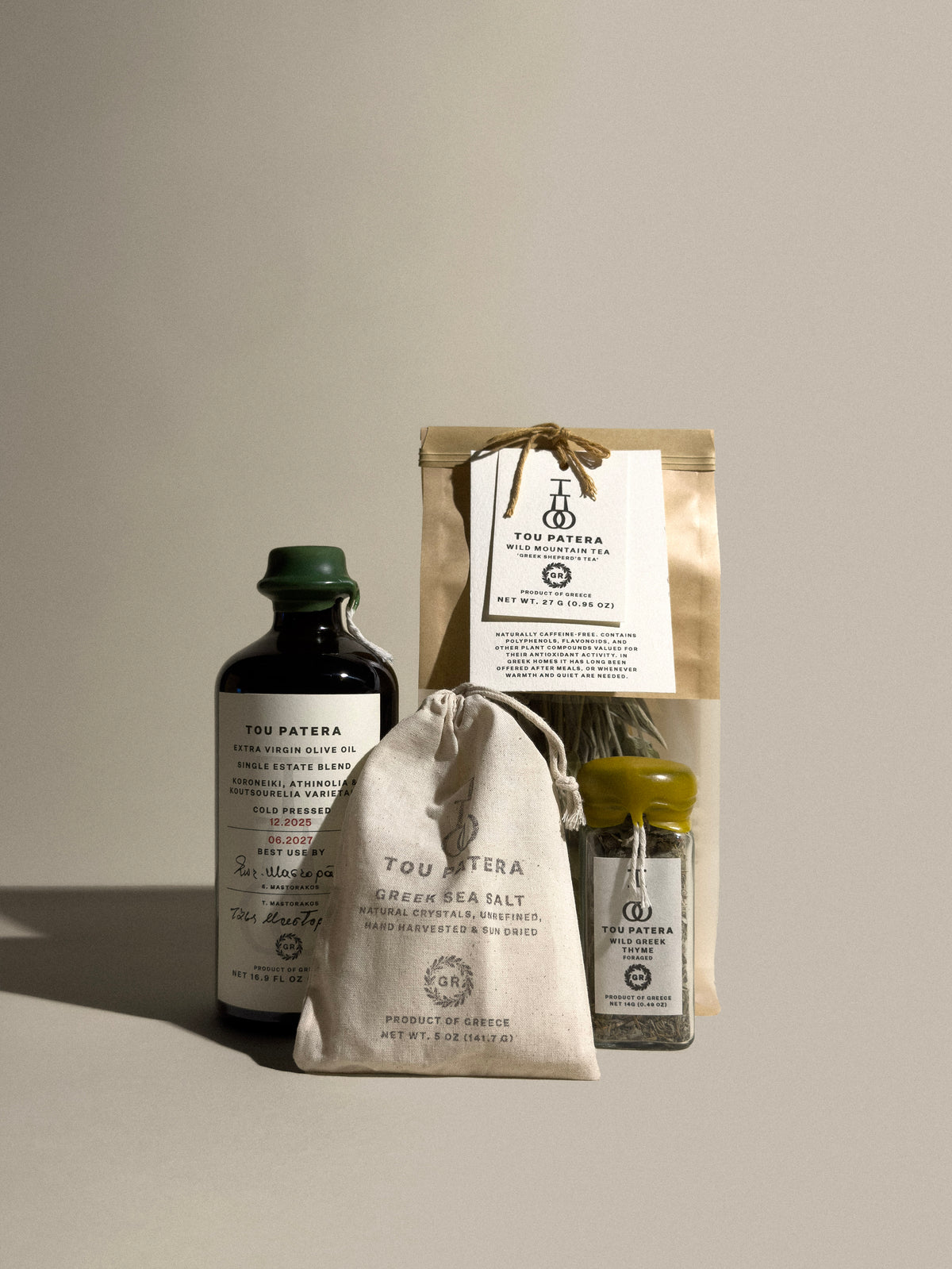 A curated product set on a warm beige background featuring a dark glass bottle of olive oil, a beige drawstring bag of sea salt, a small jar of thyme, and a kraft paper pouch, all branded Tou Patera.