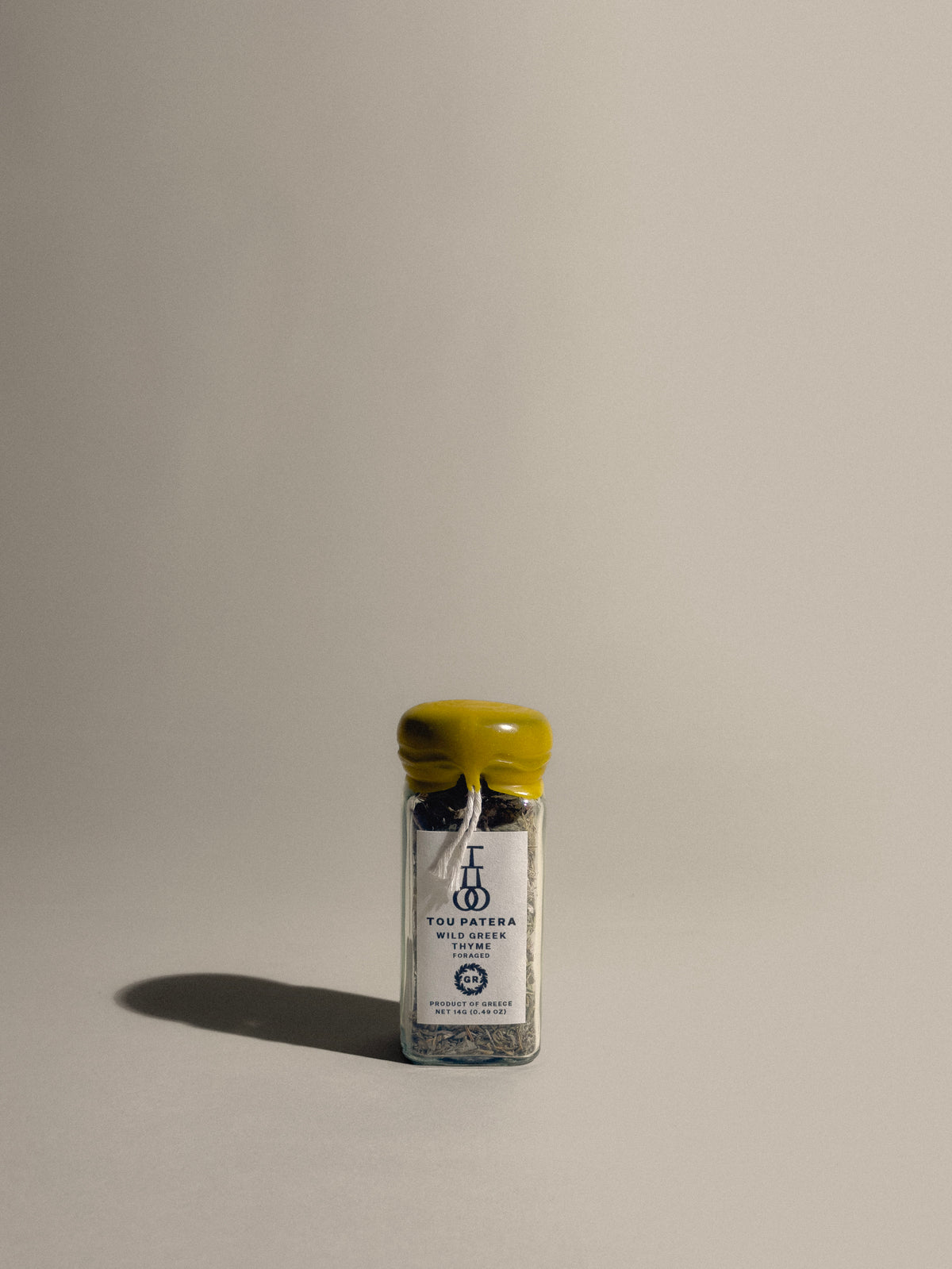 A small clear glass spice jar with a yellow lid and a white hanging label, photographed on a plain warm beige background with a soft shadow.