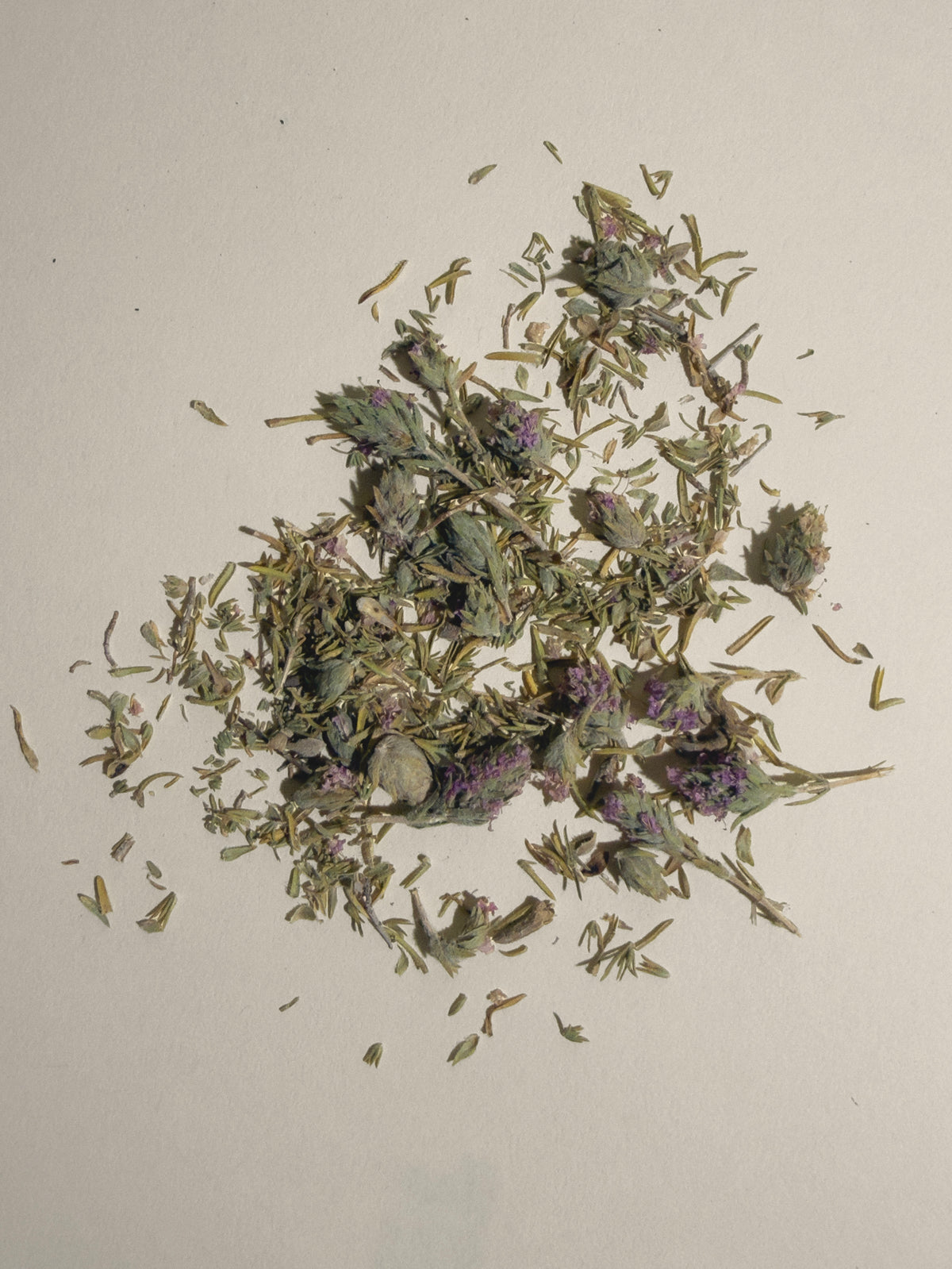 A small pile of dried loose thyme with pale green, tan, and muted purple tones, scattered on a plain off-white background.